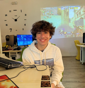 Picture of Jake Torres sitting in a computer lab with a large wall clock, projection of a video game on a while wall, and a desk with desktop computer behind him.