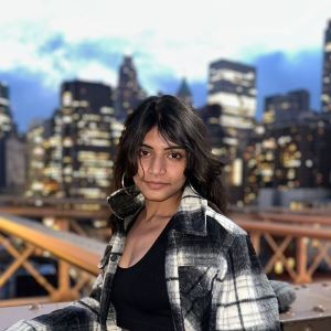 Anusha Lavanuru wearing a black and white flannel jacket in front of NYC buildings at twilight.