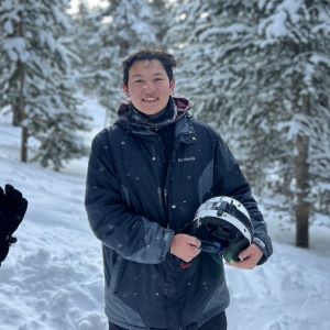 Photo of Benjamin Yang holding a snowboarding helmet and standing in the snow with snow covered trees in the background
